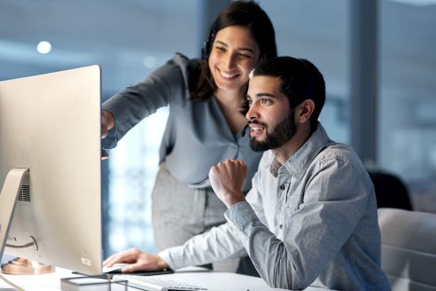 A man is sitting at a desk looking at a computer screen with a women standing next to him who is pointing to the computer screen