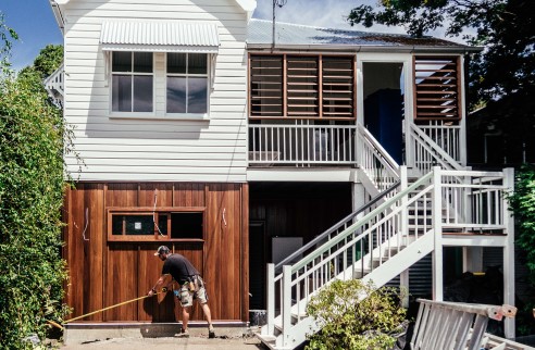 A two storey timber Queenslander house with a man sweeping the concrete driveway